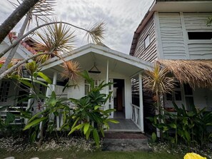 A white house with a covered porch, surrounded by tropical plants and a thatched roof. - Habana Hotel (Kuta)