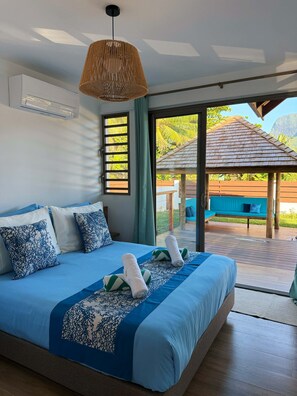 A modern bedroom with a large bed, a hanging light fixture, and a view of a wooden deck and outdoor seating area. - Perfect Moorea Getaway for Families & Friends – Pool + Beach 200m (Moorea)