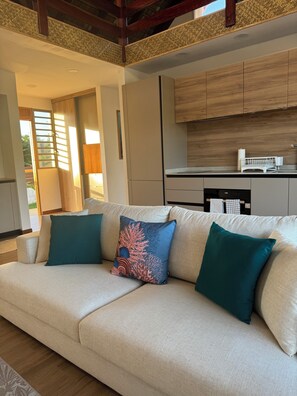 A modern living room with a beige sofa, teal cushions, and a kitchen area with wooden cabinets and a dishwasher. - Perfect Moorea Getaway for Families & Friends – Pool + Beach 200m (Moorea)