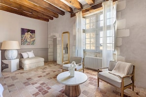 Une pièce avec un sol à carreaux, une table ronde en bois, un canapé blanc, une chaise en bois, un grand miroir et un tableau accroché au mur.
