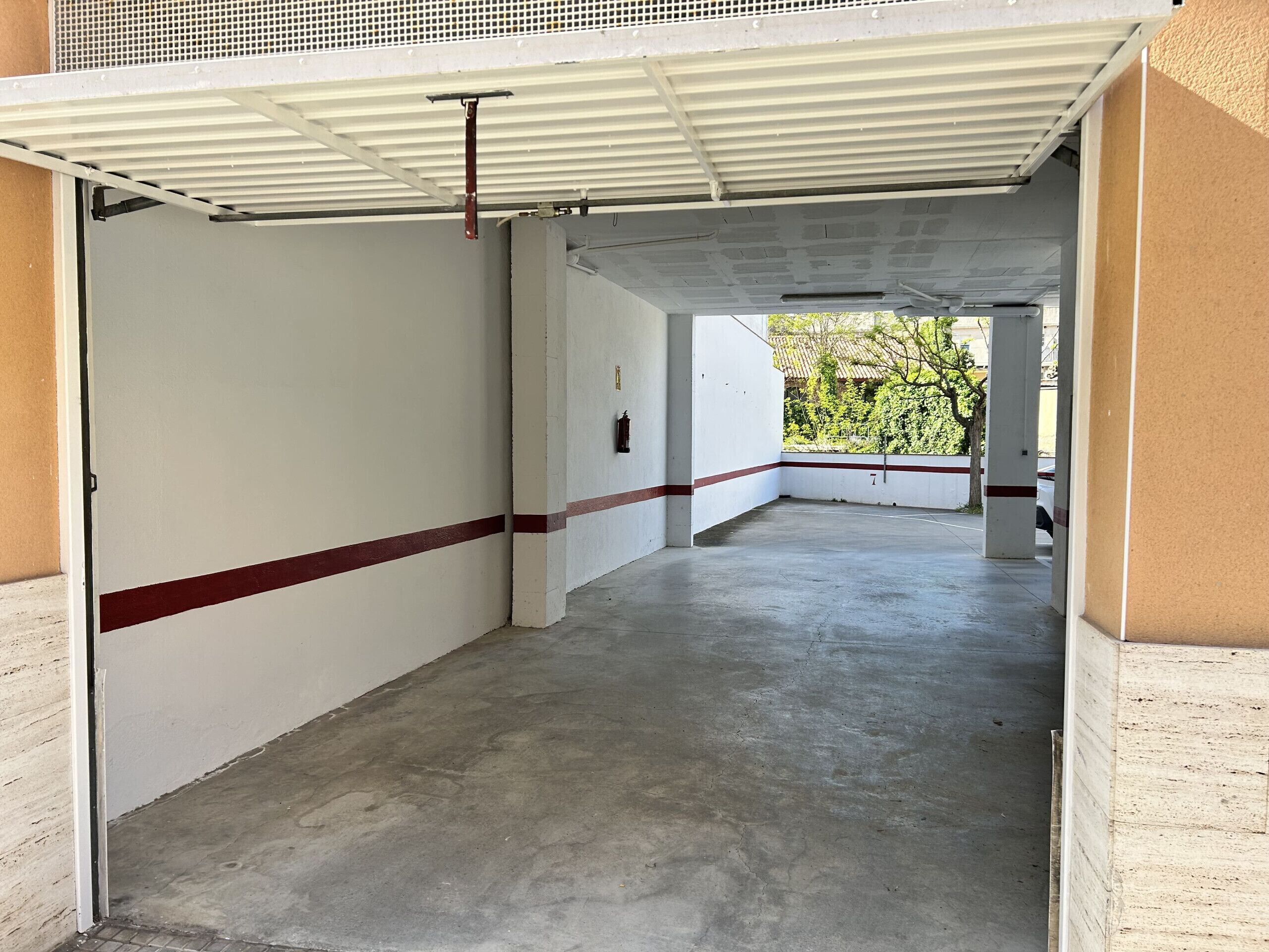 A corridor with a white wall and red stripe, leading to a covered area with a metal roof.