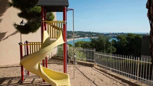 Un'area giochi con uno scivolo giallo e una struttura di arrampicata rossa, con vista su una spiaggia e una località costiera.