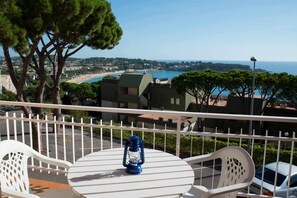 Un balcone con tavolo e sedie bianche, affacciato su una cittadina costiera con edifici e una spiaggia.