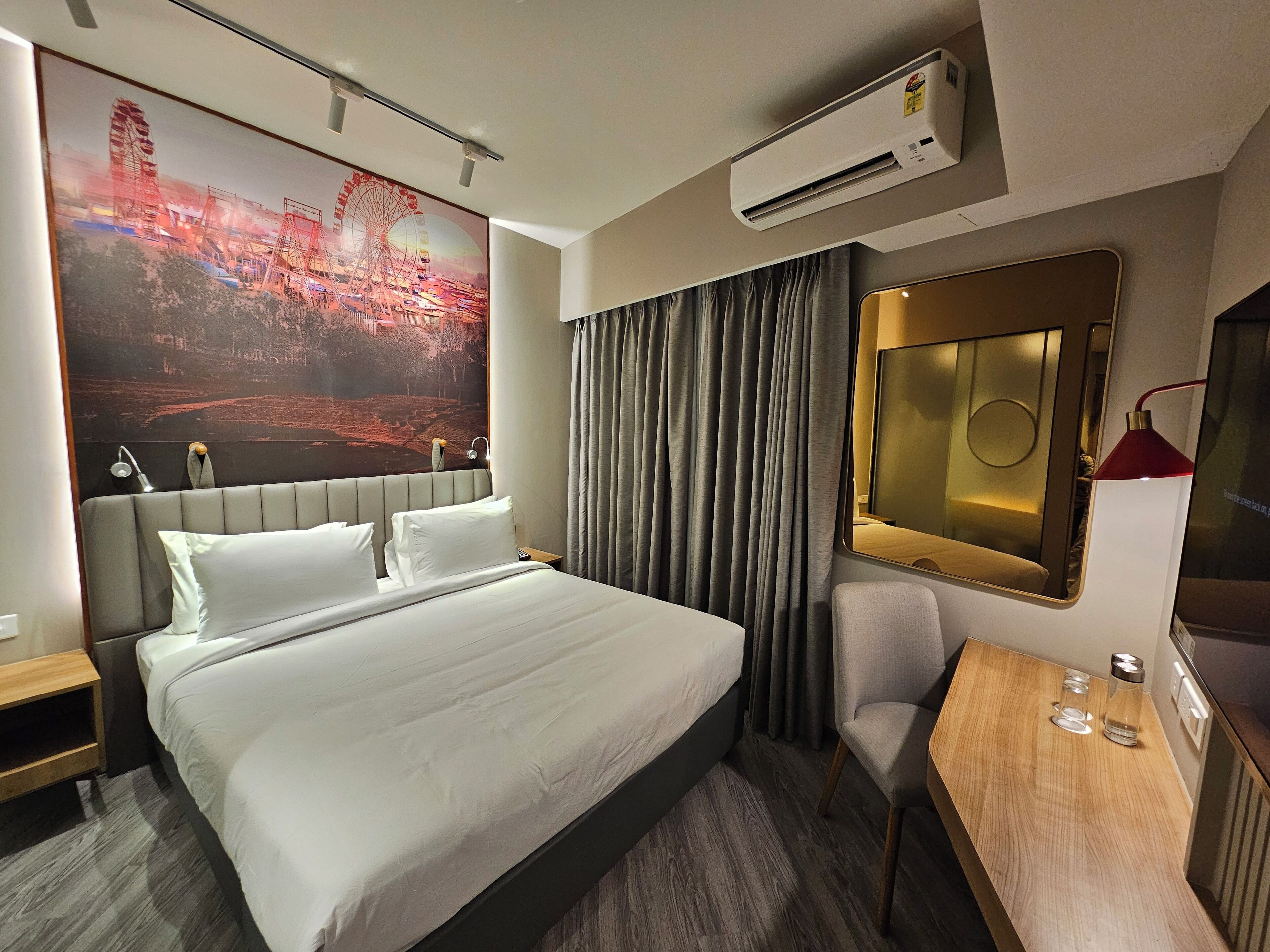 A hotel room with a large bed, a desk, a chair, an air conditioner, and a large poster on the wall.