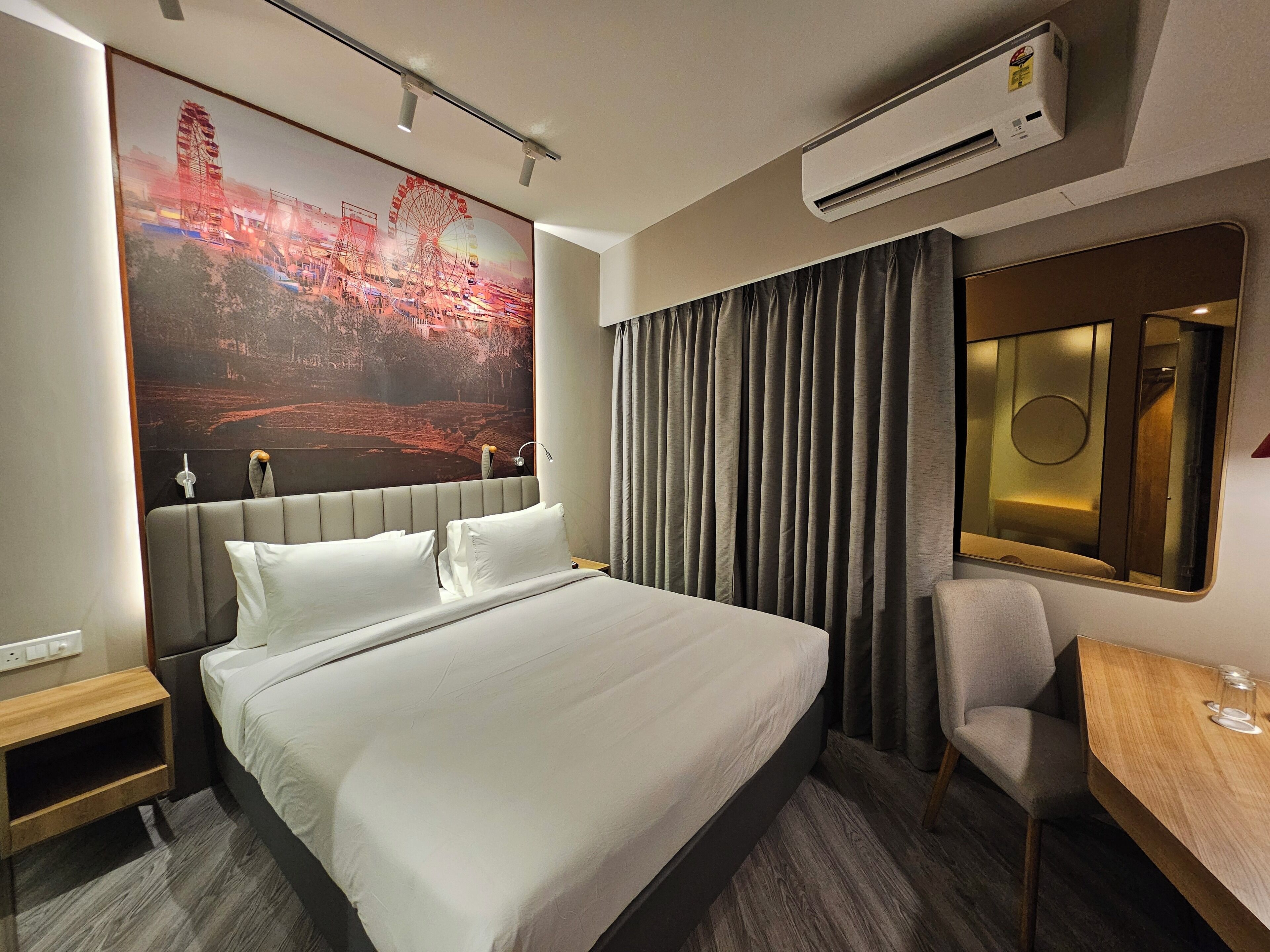 A hotel room with a large bed, a desk with a chair, an air conditioner, and a large poster on the wall.