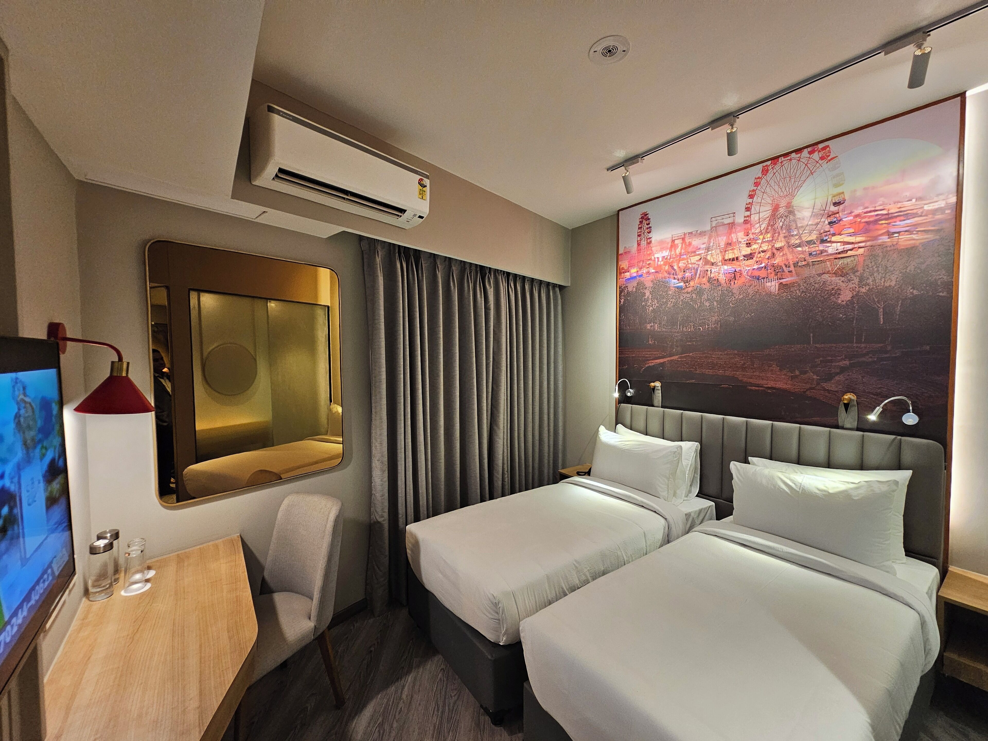 A hotel room with two beds, a desk with a computer, a mirror, an air conditioner, and a large window with a cityscape view.