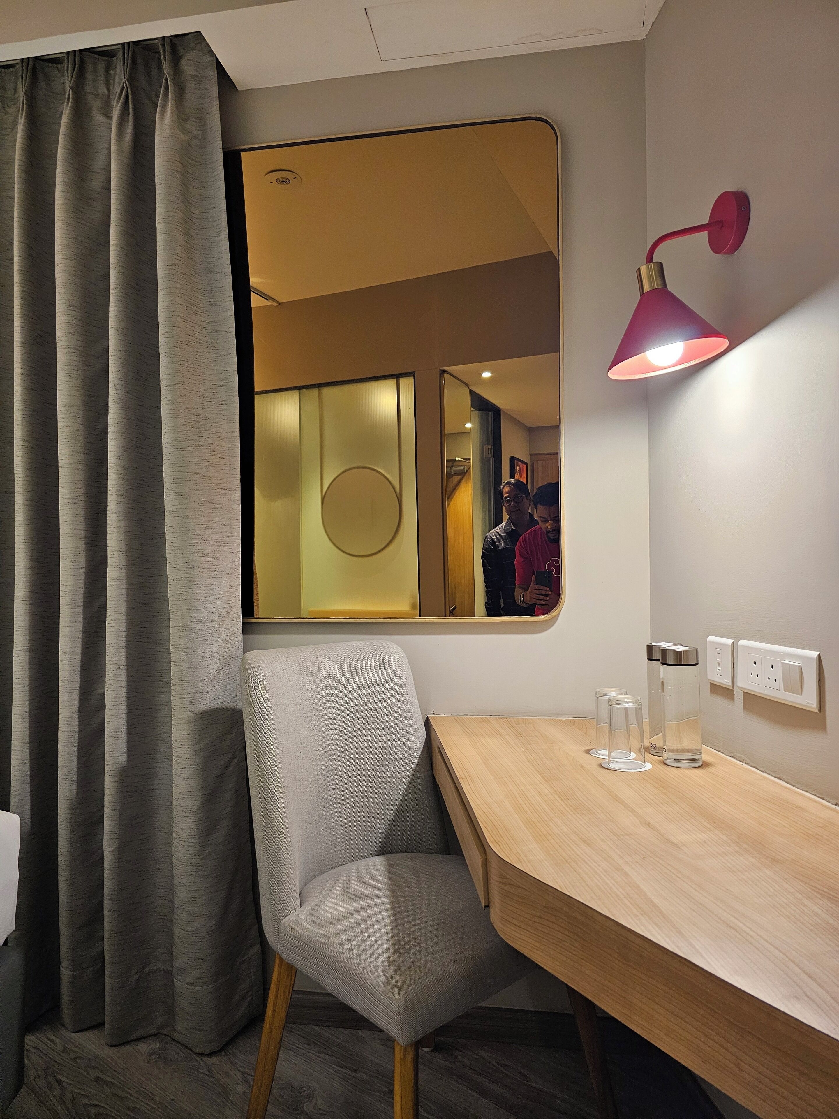 Habitación de hotel con un escritorio de madera, una silla gris, un espejo, una lámpara roja y una ventana con cortinas grises.