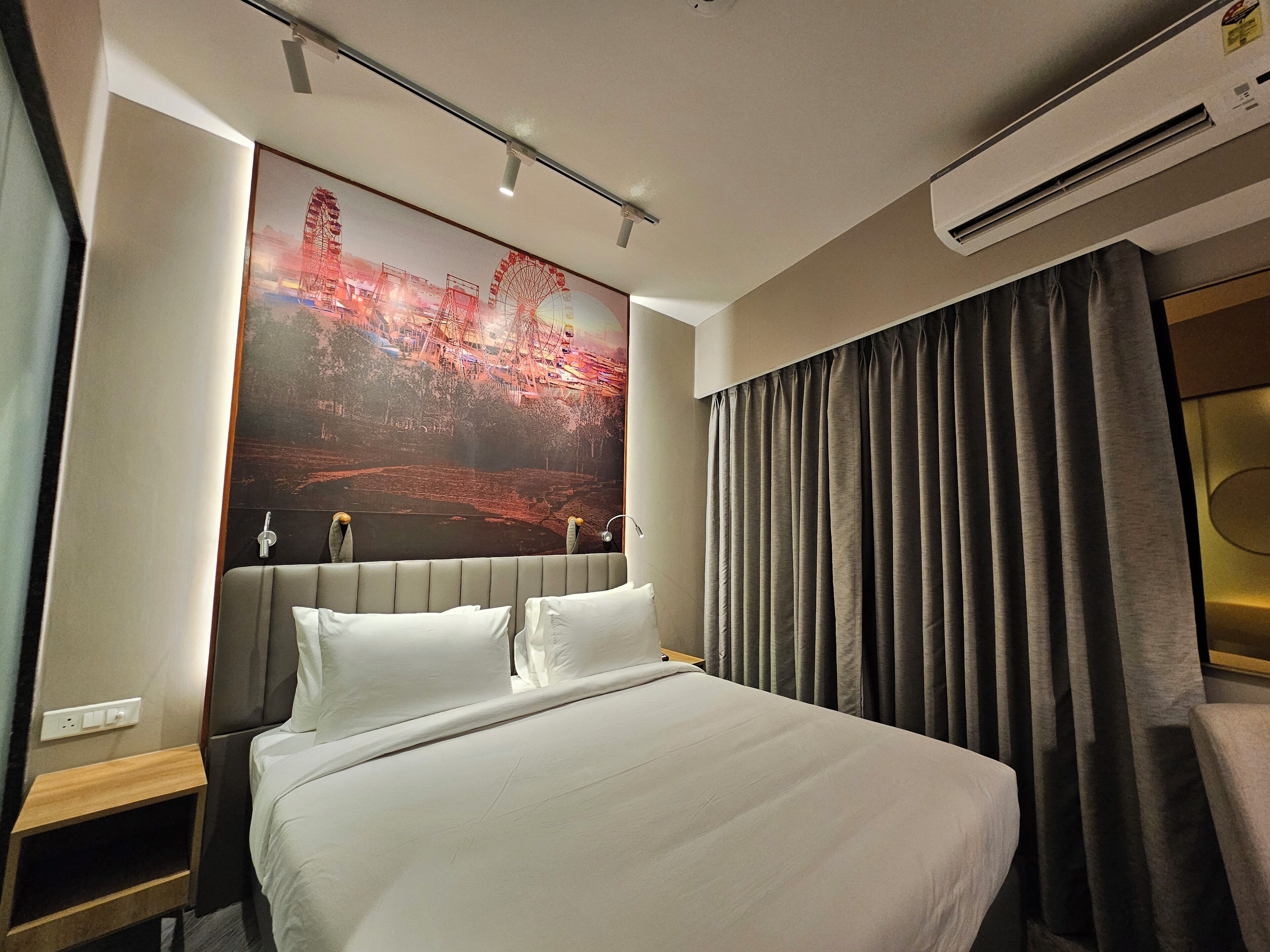 A hotel room with a large bed, a headboard with a cityscape mural, a bedside table, and a window with curtains.