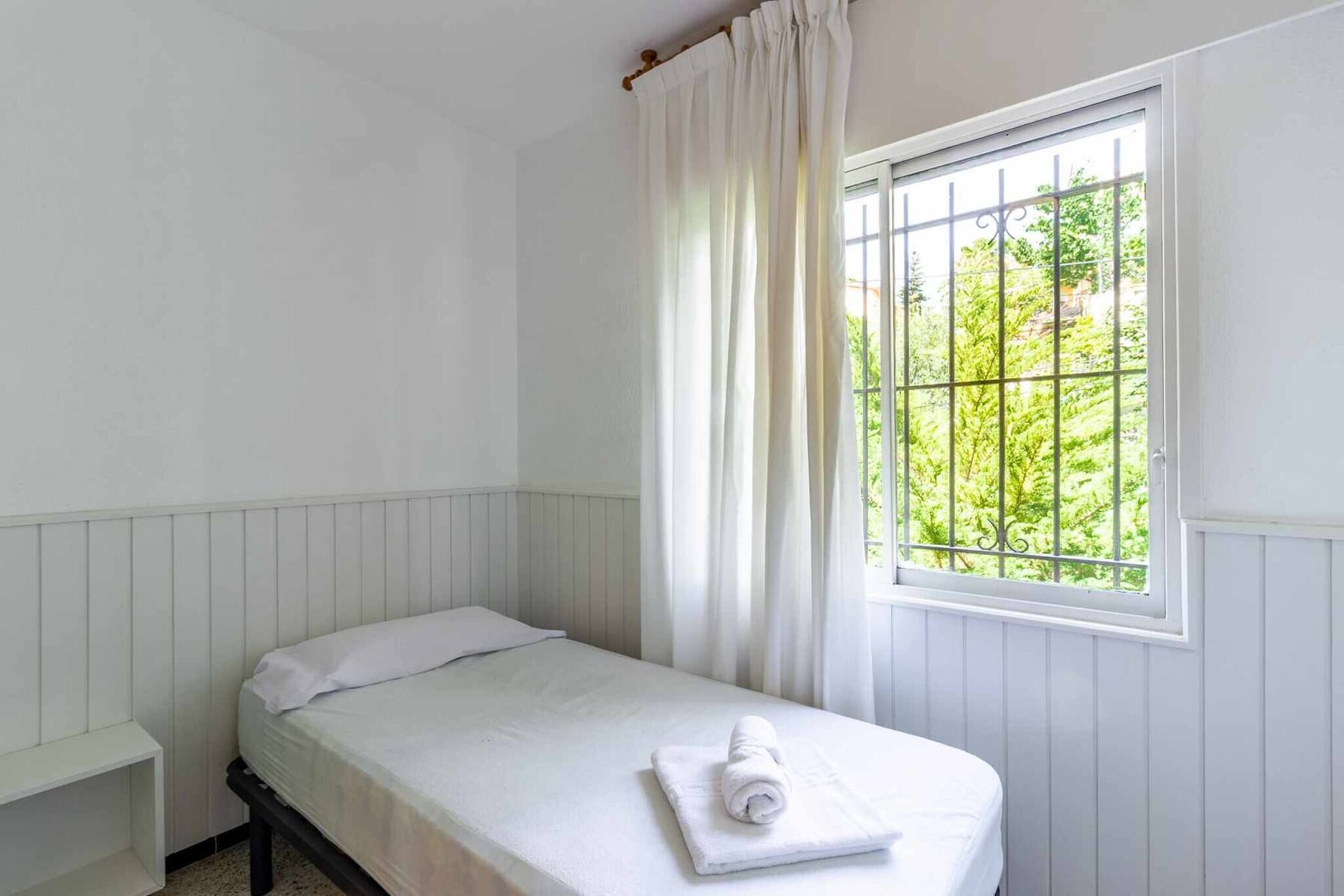 Een kleine, schone kamer met een enkel bed, een raam met metalen tralies en uitzicht op groen.
