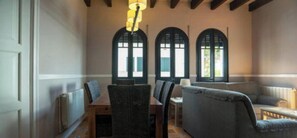 Ein Essbereich mit Tisch, Stühlen und einer Couch. Drei Bogenfenster mit Fensterläden sind vorhanden.