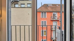 Une vue à travers une porte donnant sur un balcon, un immeuble à la façade rouge et un ciel dégagé.