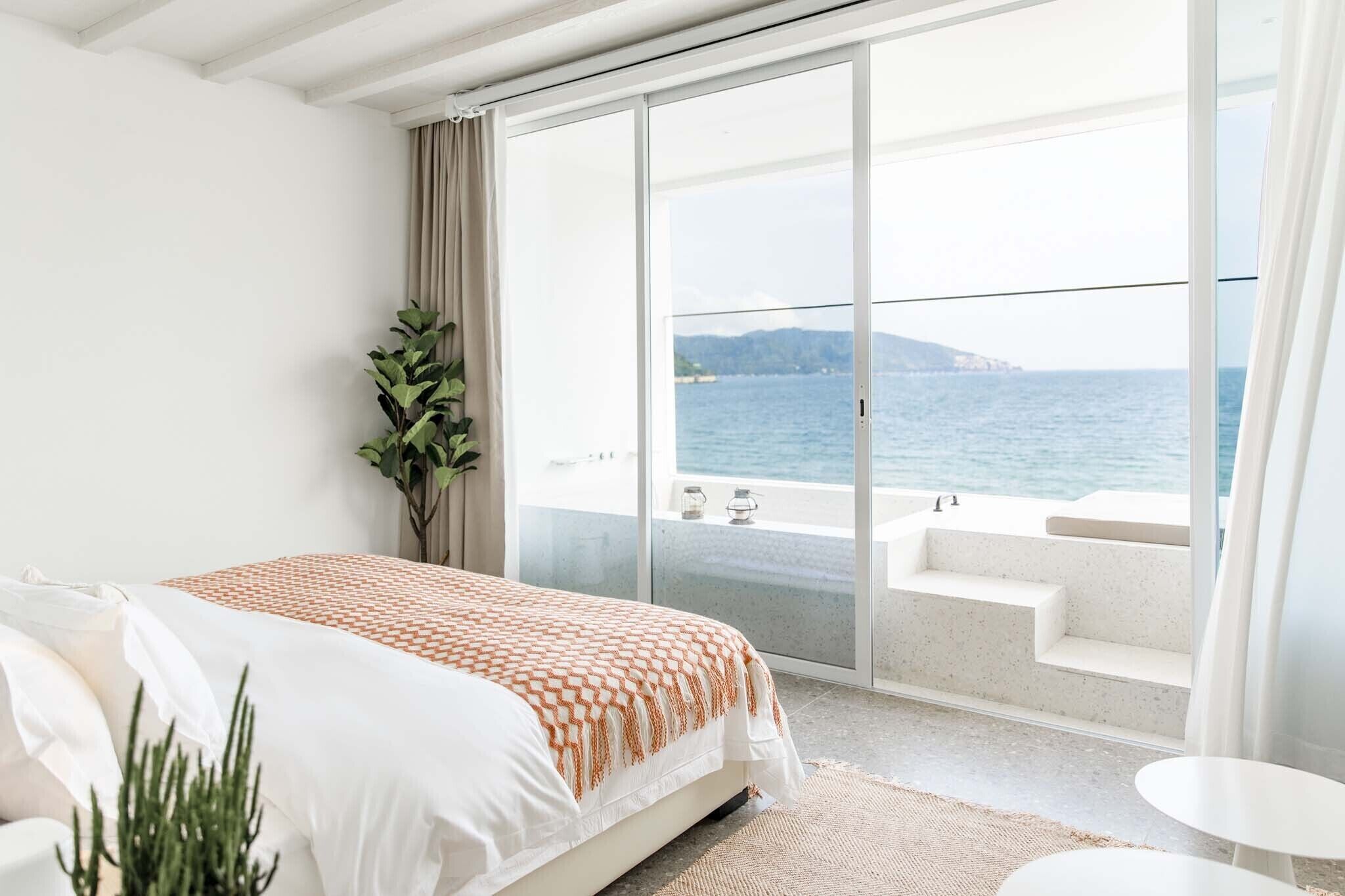 Un dormitorio con una cama grande, una planta en maceta y vistas al mar a través de una puerta corrediza de vidrio.