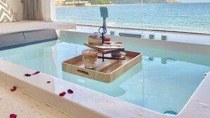 Una bañera con pétalos de rosa, una bandeja con libros y un vaso de agua, y vista al mar y las colinas.