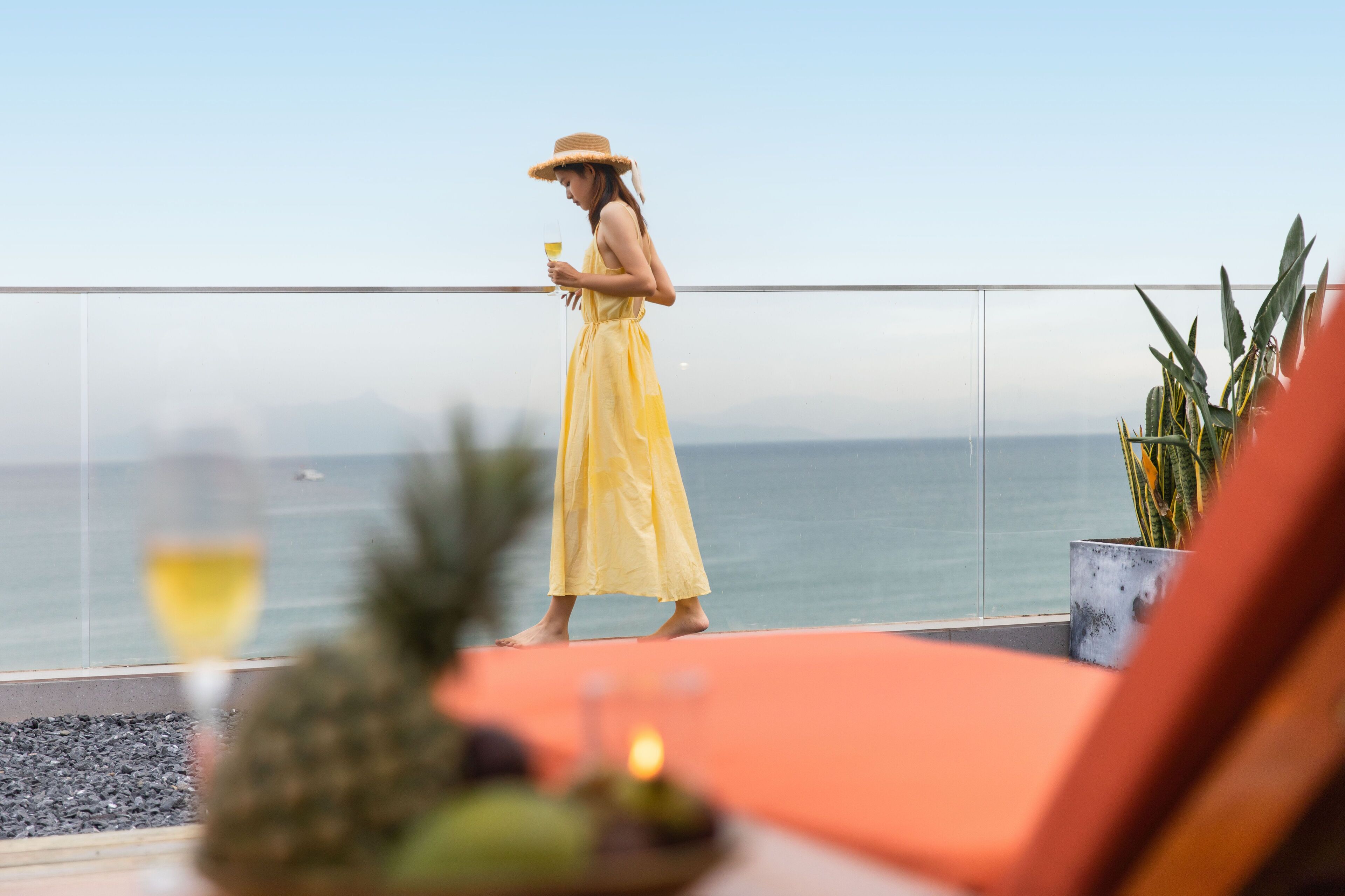 A person in a yellow dress standing on a balcony overlooking the sea, with a potted plant and a glass of drink on the table.