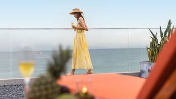 A person in a yellow dress standing on a balcony overlooking the sea, with a potted plant and a glass of drink on the table.