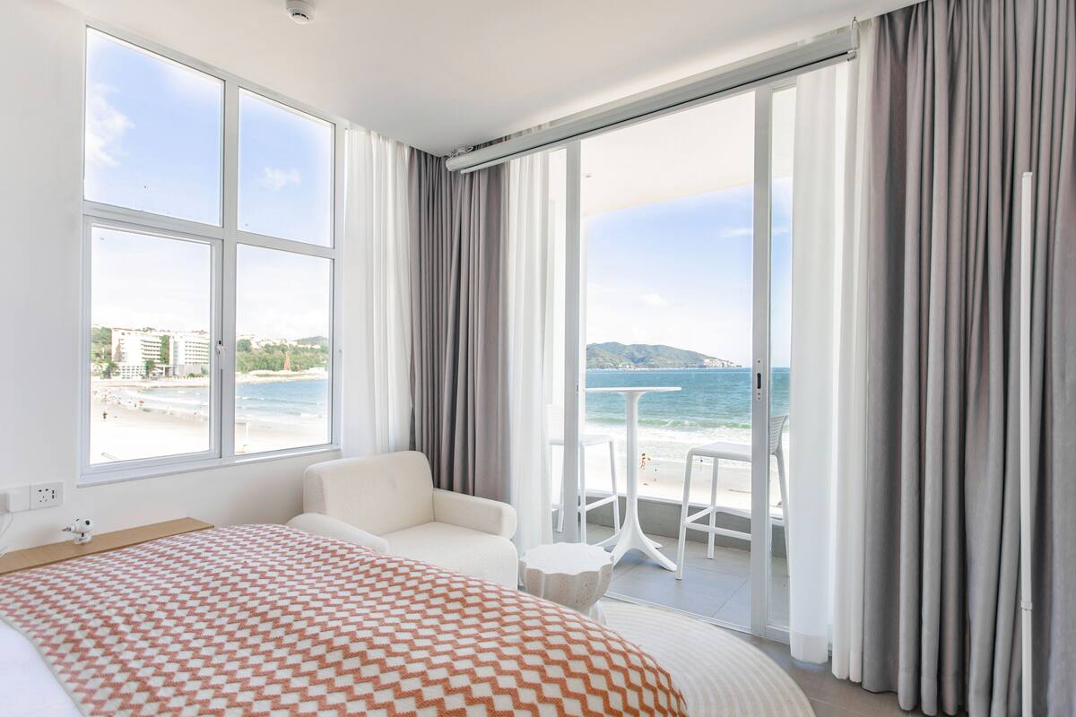 A hotel room with a large bed, a white armchair, a small round table, and a balcony with a view of the beach and sea.
