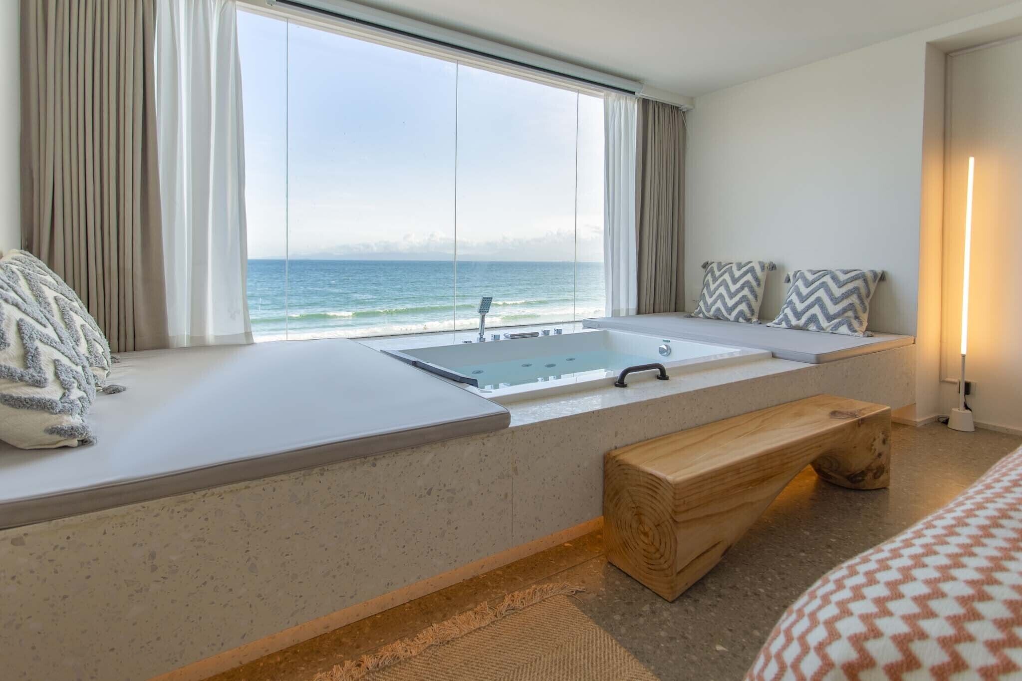 Une chambre d’hôtel dotée d’une grande fenêtre donnant sur la mer, d’une baignoire intégrée, d’un banc en bois et d’un lit avec une literie à motifs.
