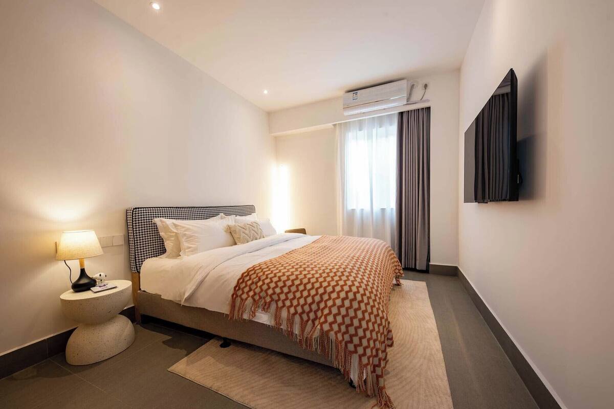 A modern hotel room with a bed, a bedside lamp, an air conditioning unit, and a flat-screen TV mounted on the wall.