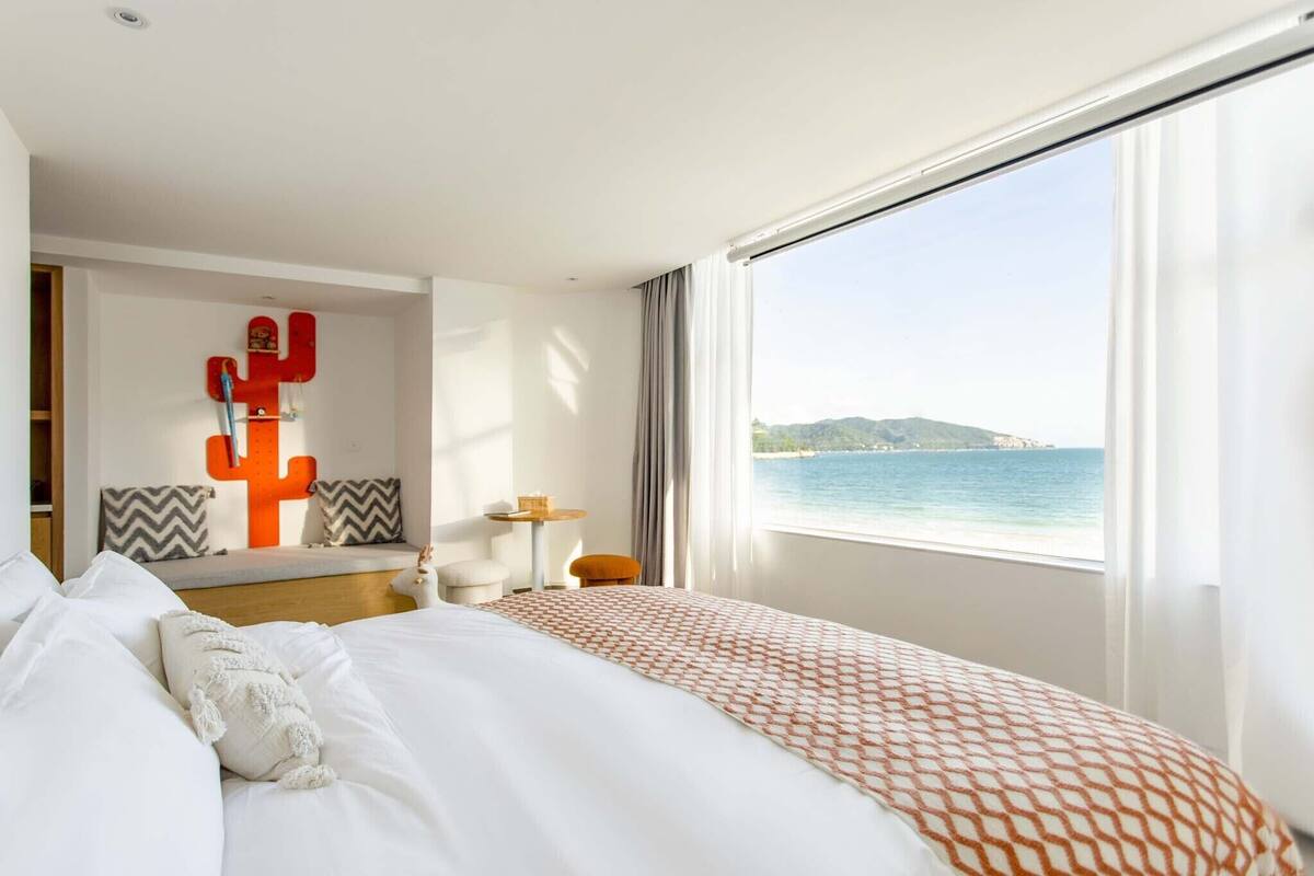 A hotel room with a bed, a cactus decoration, a view of the sea, and a window with curtains.