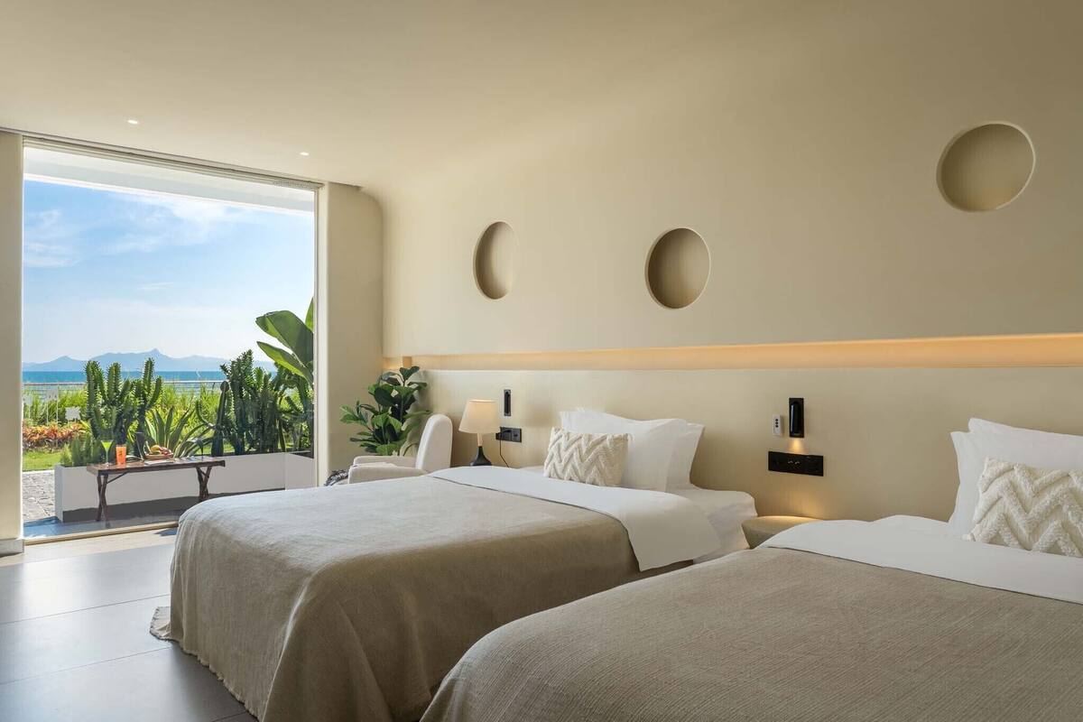 A hotel room with two beds, a large window with a view of the sea and mountains, and a small table with a lamp.