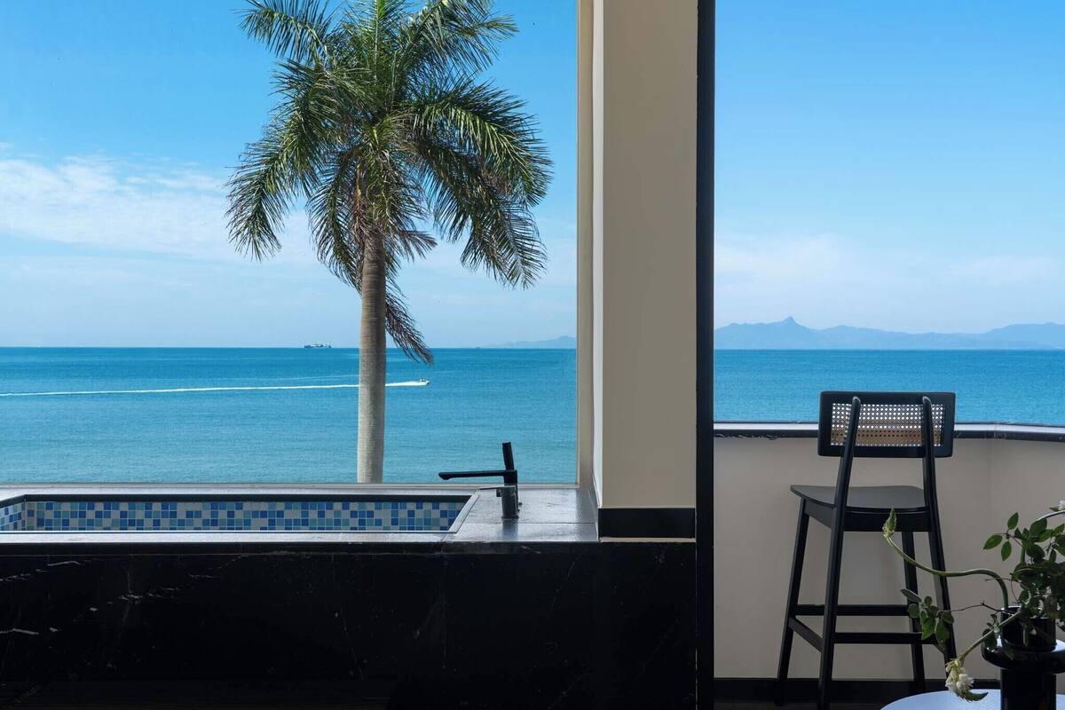 A balcony with a view of the sea, a palm tree, a pool, and a chair.