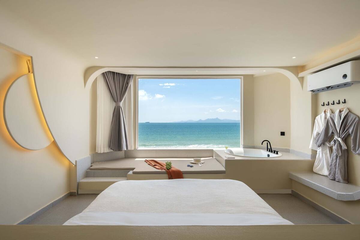 A modern hotel room with a large bed, a view of the sea, a bathtub, and a robe drying rack.