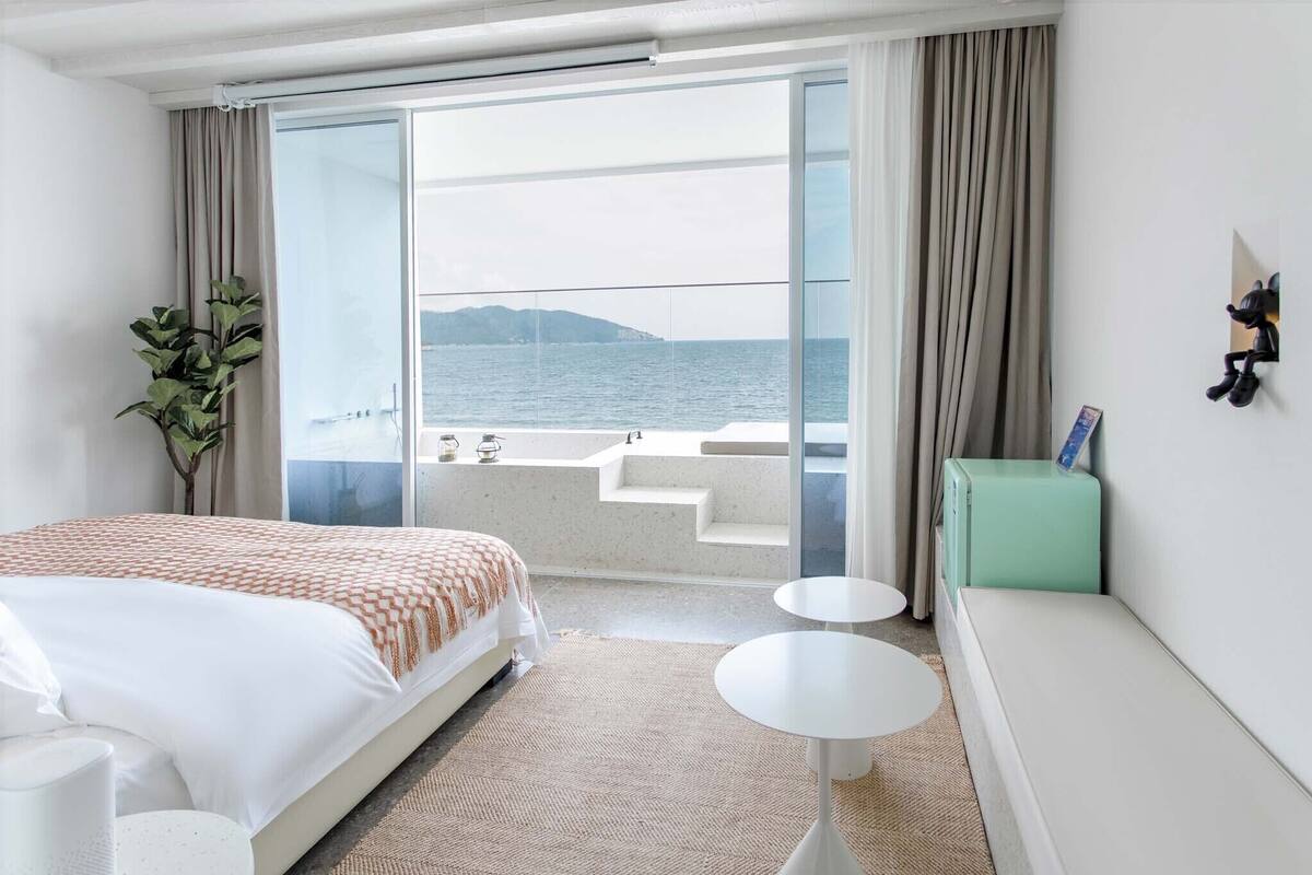 A hotel room with a bed, a small table, a sofa, a plant, and a view of the sea through a sliding glass door.