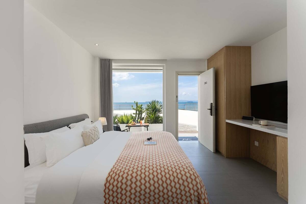A hotel room with a bed, a TV, a balcony with a view of the beach, and a door leading outside.