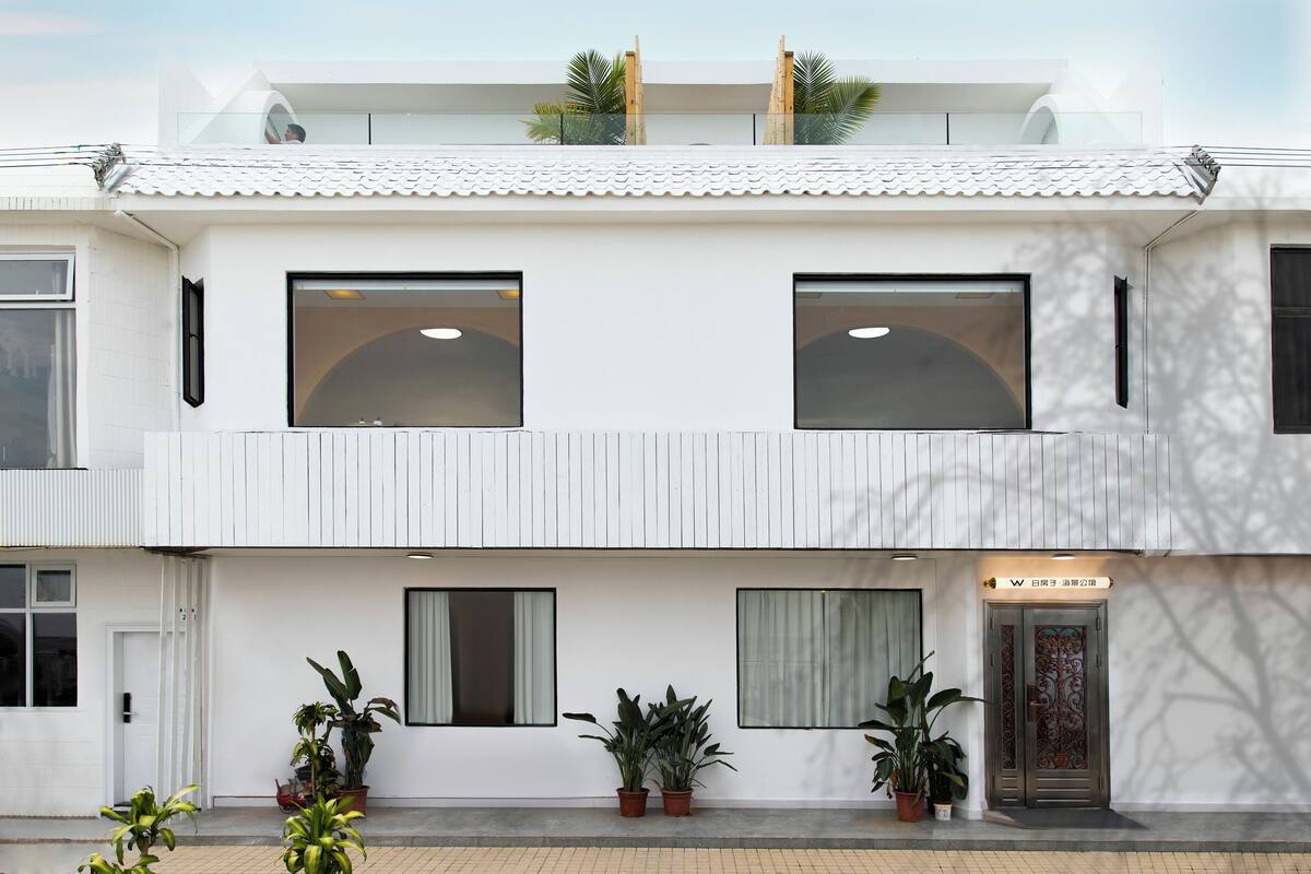 A modern building with a white facade, multiple windows, and a balcony with plants.