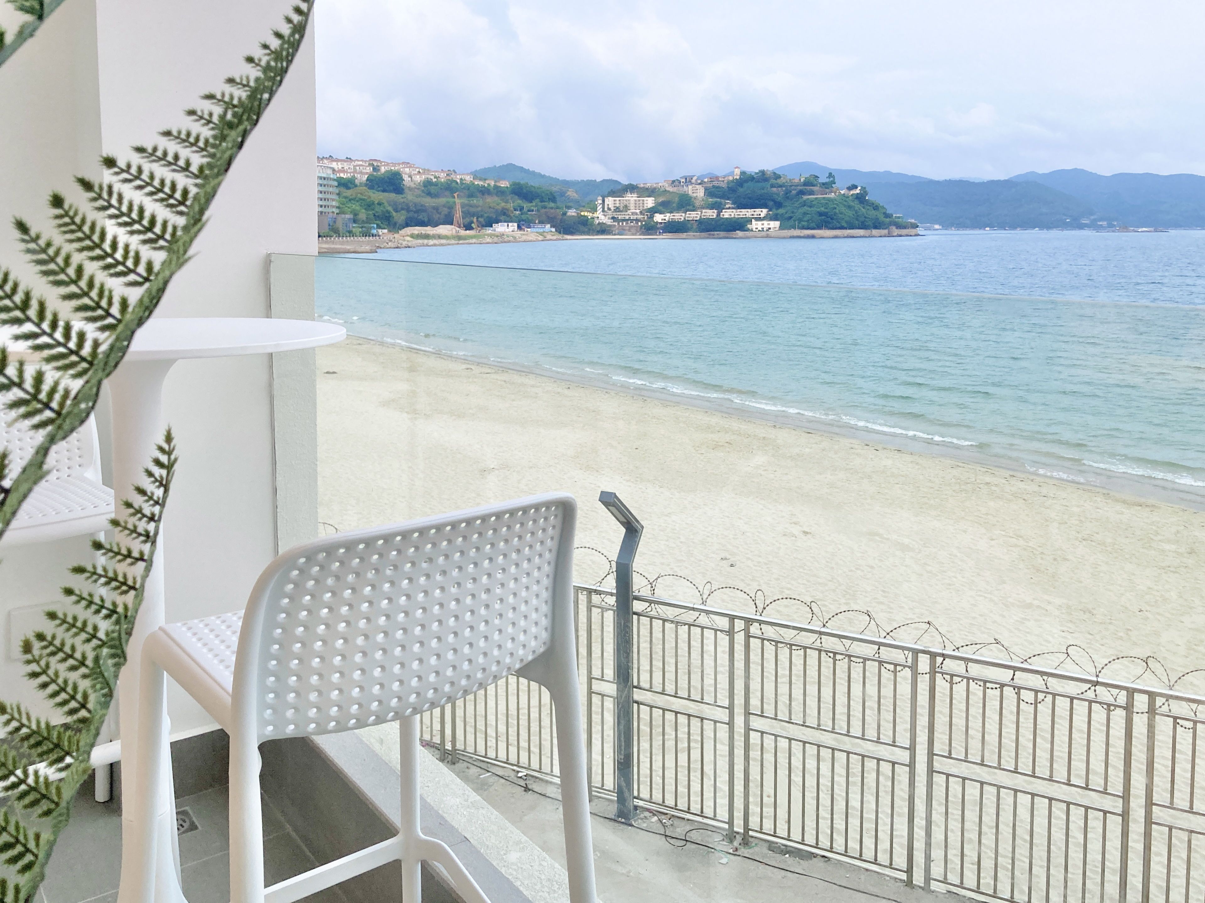 A balcony with a white chair and a small table, overlooking a beach with clear water and distant hills.