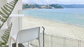A balcony with a white chair and a small table, overlooking a beach with clear water and distant hills.