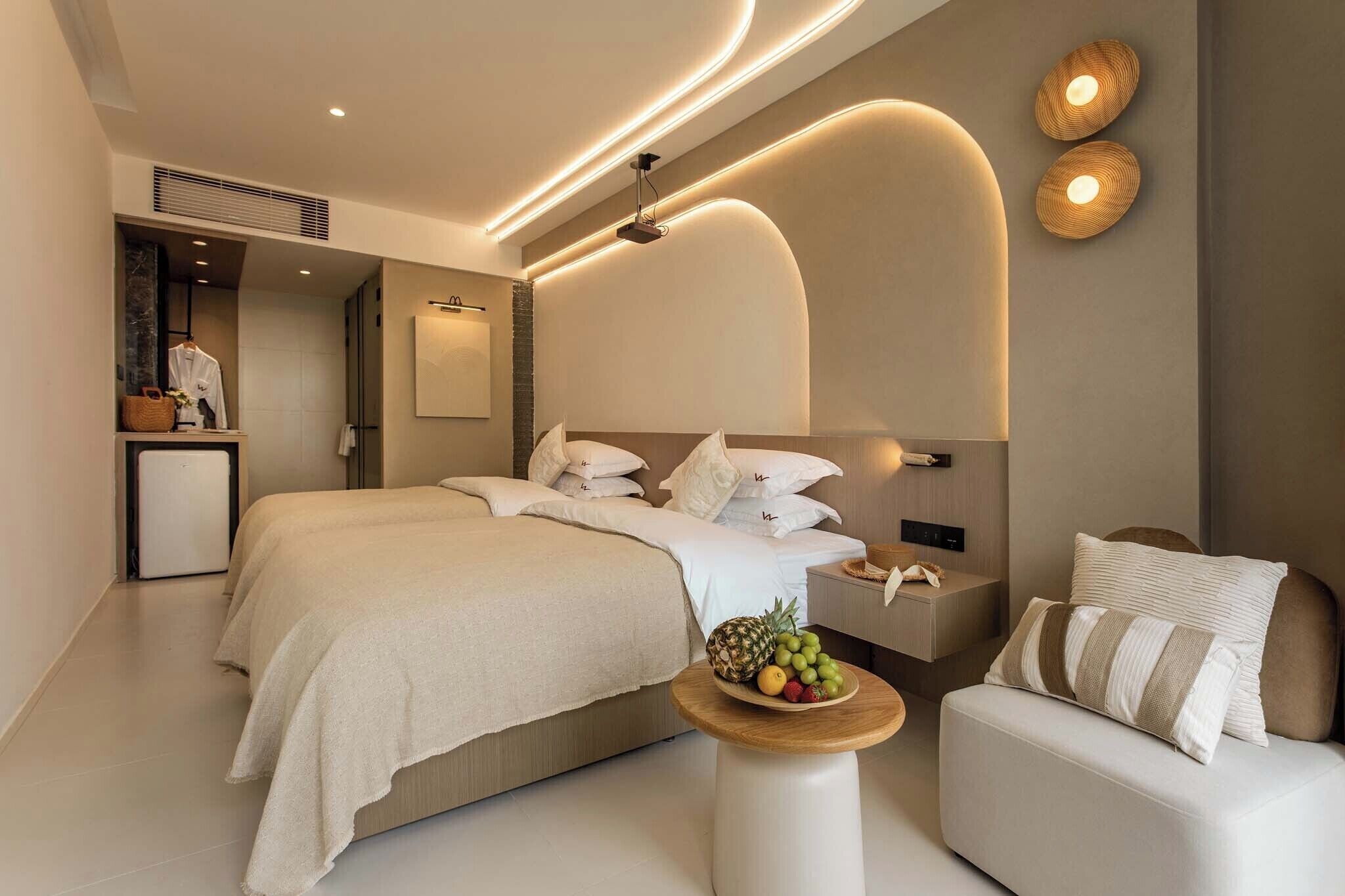 Una habitación de hotel moderna con una cama grande, mesitas de noche, un pequeño rincón de descanso y un frutero sobre una mesa redonda.