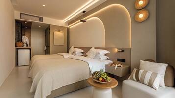 Una habitación de hotel moderna con una cama grande, mesitas de noche, un pequeño rincón de descanso y un frutero sobre una mesa redonda.