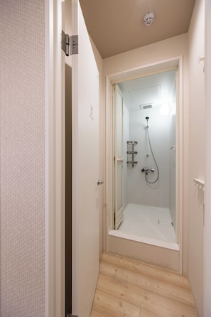 Un pasillo estrecho que conduce a una zona de ducha con puerta de cristal, una lámpara en el techo y un calentador de agua a la vista.
