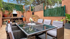 Eine Terrasse im Freien mit einem Glastisch, Korbstühlen und einem Grill.