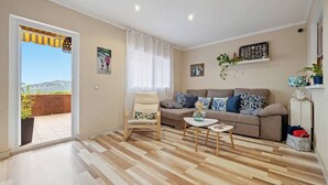 Ein Wohnzimmer mit Sofa, Sessel und Couchtisch. Ein Fenster mit Vorhängen, eine Tür zum Balkon und ein Blick ins Freie.