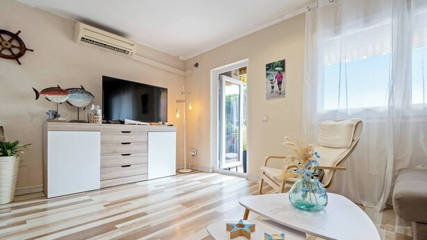 Ein modernes Wohnzimmer mit Fernseher, Holzboden, einem weißen Sofa, einem Esstisch mit Vase und einem Stuhl.