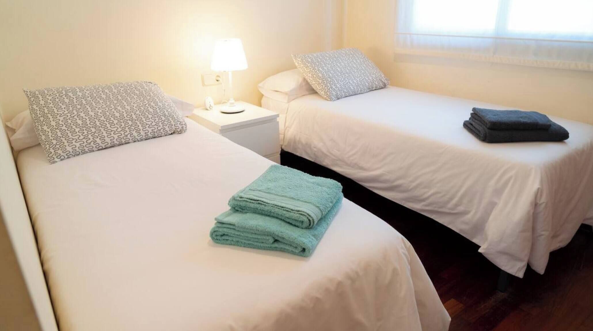 A hotel room with two single beds, each with a pillow and a folded towel. There is a nightstand with a lamp and a phone.