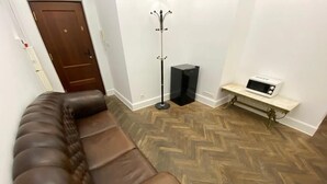 Una habitación con sofá de cuero marrón, suelo de madera tipo espiga, perchero, altavoz negro y un electrodoméstico blanco pequeño sobre una encimera de mármol.
