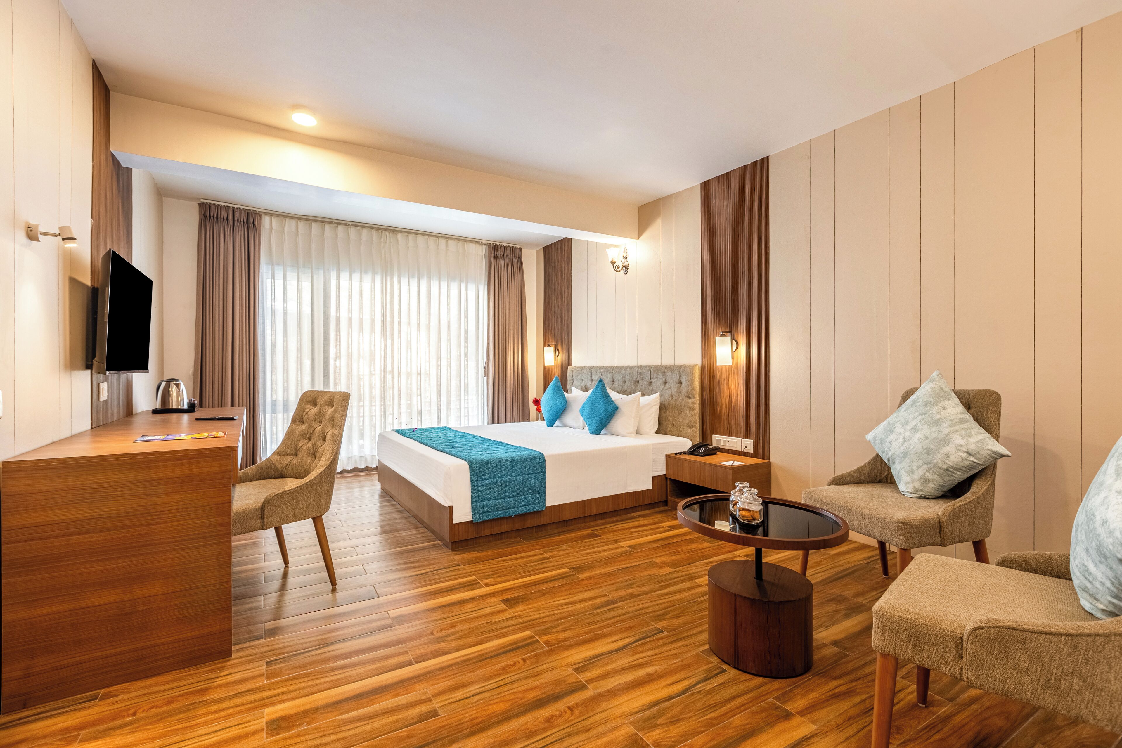 A modern hotel room with a wooden floor, a bed with blue and white bedding, a desk with a flat-screen TV, a coffee table, and two armchairs.