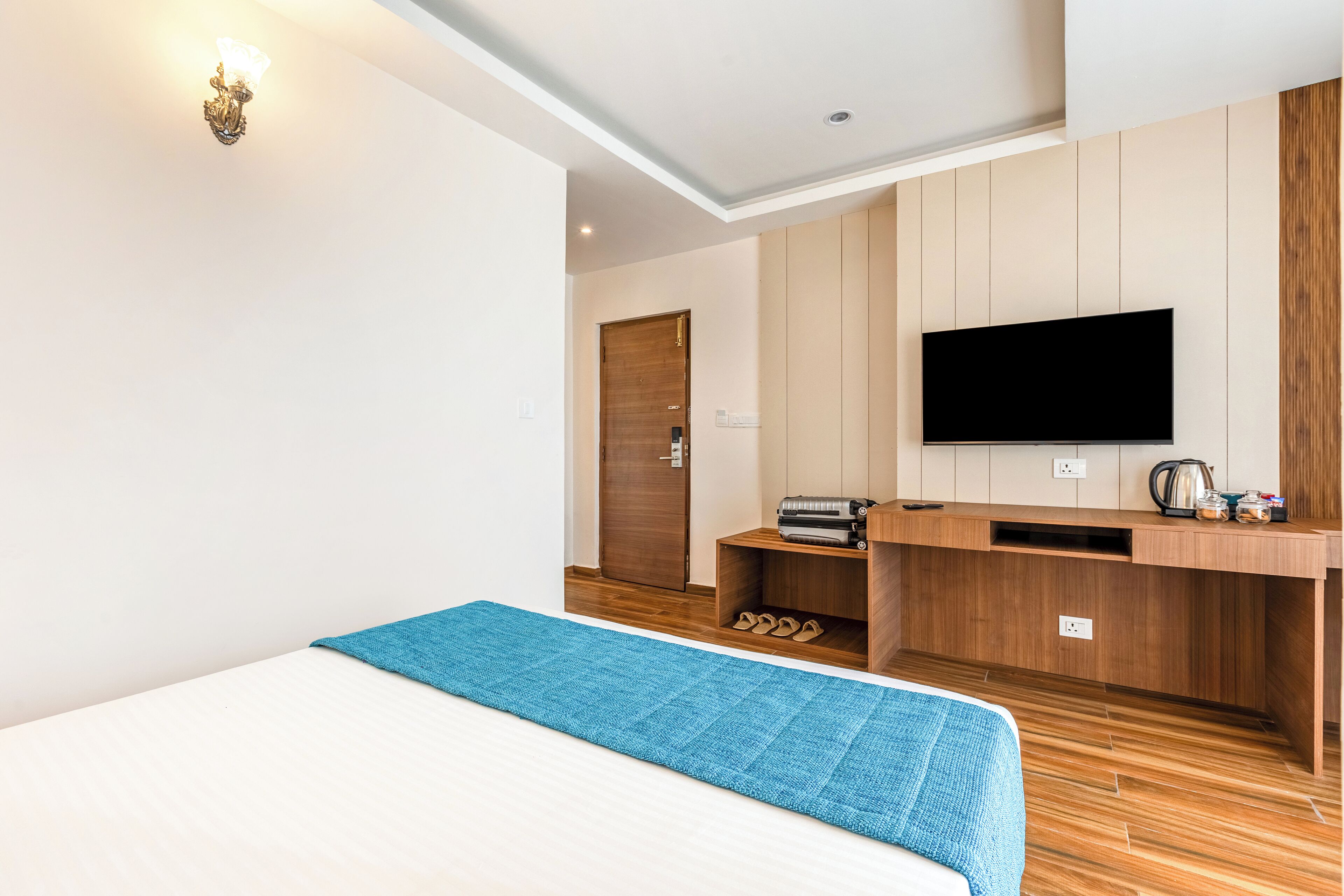A modern hotel room with a bed, a TV mounted on the wall, a wooden desk, and a shoe rack.