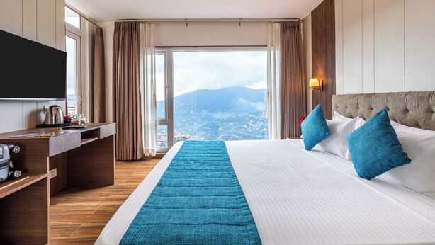 A hotel room with a bed, a desk, a flat-screen TV, and a view of mountains through a large window. - Sterling Mount Olive Gangtok (Gangtok)