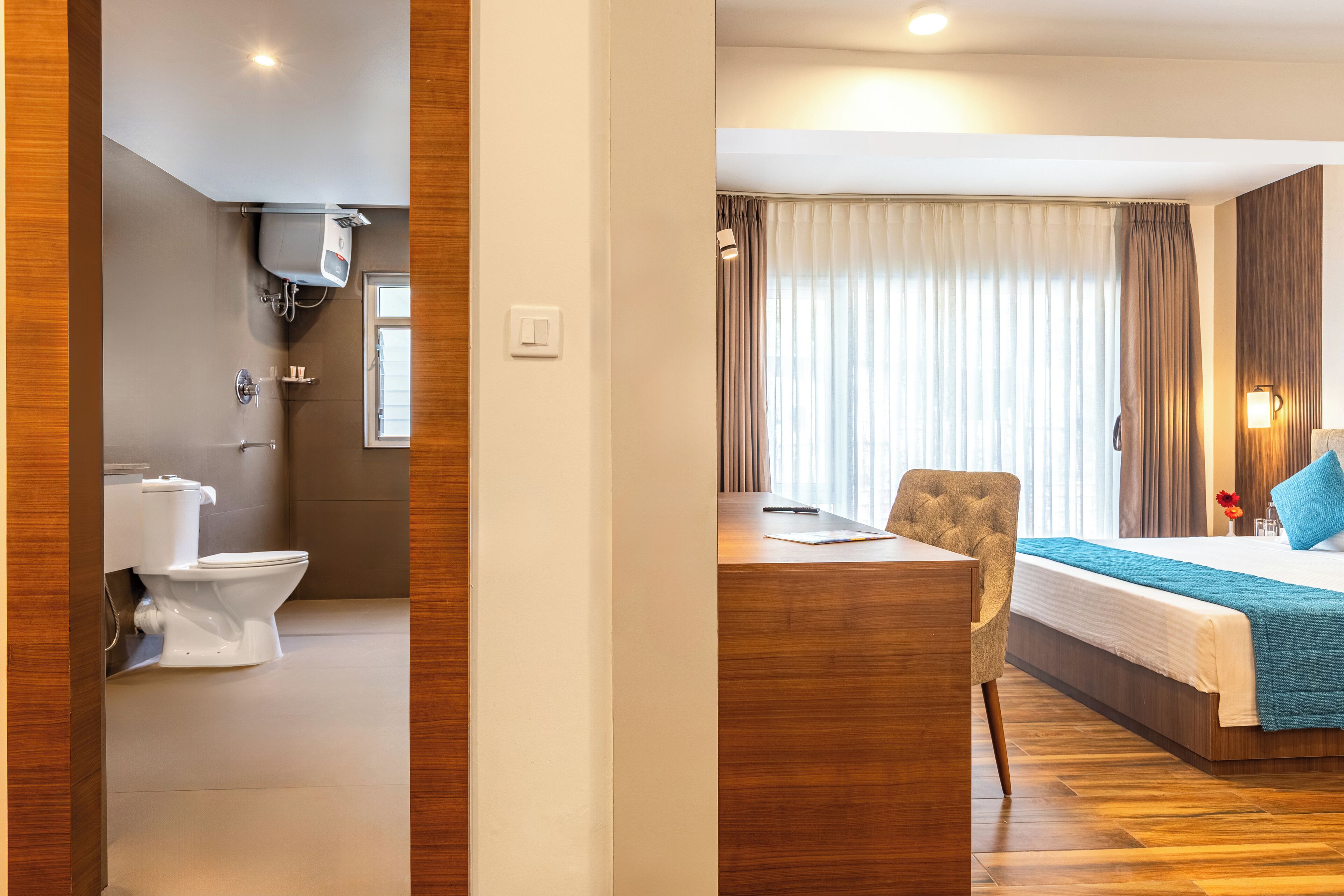 Una camera d'albergo con pavimento in legno, un letto con coperta blu, una scrivania in legno, una sedia, un bagno con WC e doccia, e una finestra con tende.