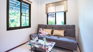A modern living room with a grey sofa, a glass coffee table with magazines, a vase with flowers, and two windows with blinds. - Oxavia Garden Bungalow B1-B2 (Rawai)
