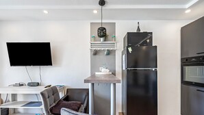 Ein kompakter Wohnbereich mit Flachbildfernseher, einem Essbereich mit Tisch und Stühlen, einer integrierten Küche mit Kühlschrank und Backofen sowie einer Pendelleuchte.