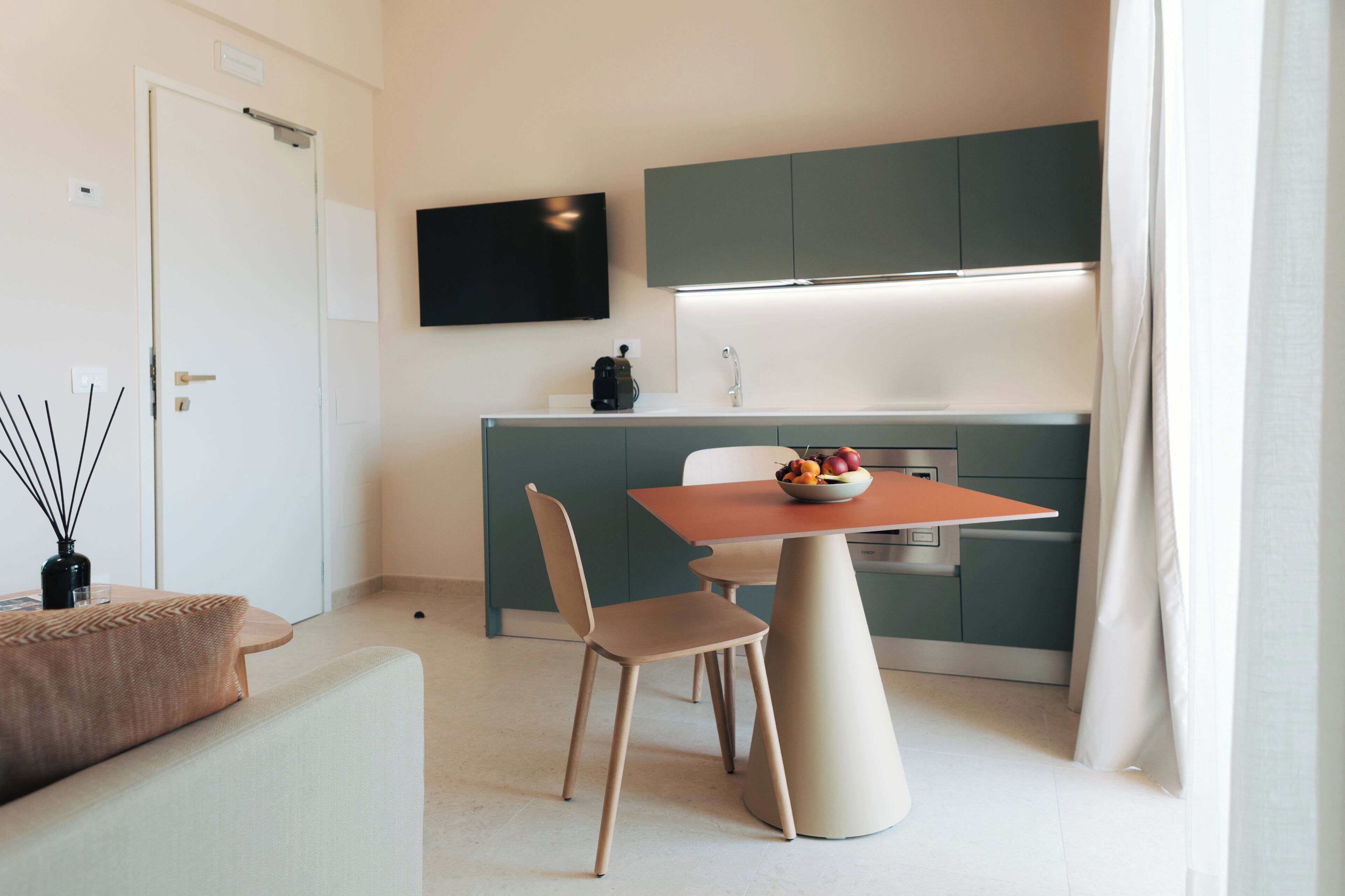 A compact living space with a kitchenette, a dining table with chairs, a flat-screen TV mounted above the counter, and a small seating area with a sofa and a coffee table.