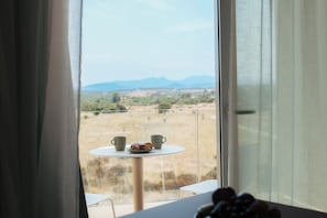 A room with a view of a field and mountains through a window, a table with two cups and a plate, and a small plant on the windowsill. - Palazzo Rubino ApartHotel (Olbia)