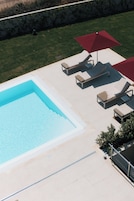Una piscina con sillones de descanso y sombrillas, rodeada por un muro de piedra y una zona verde.