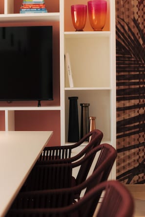 Une pièce avec un téléviseur posé sur un meuble, une table blanche et une bibliothèque contenant des livres et des objets décoratifs.