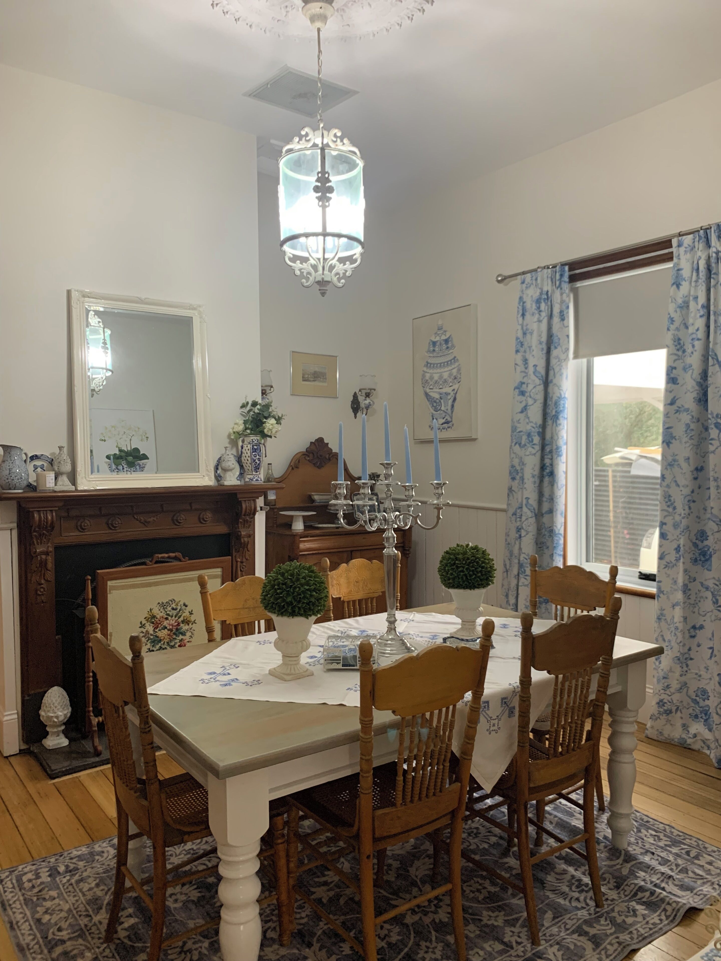 Uma sala de jantar com um candelabro, cadeiras de madeira, uma mesa com toalha de mesa florida e uma lareira.